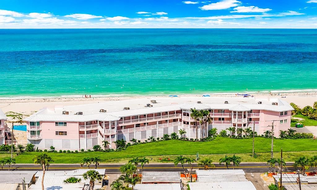 Close aerial of the pink-trimmed Anna Maria Island Club building from the Gulf side, with white sand beach in the mid-ground and turquoise water across the foreground